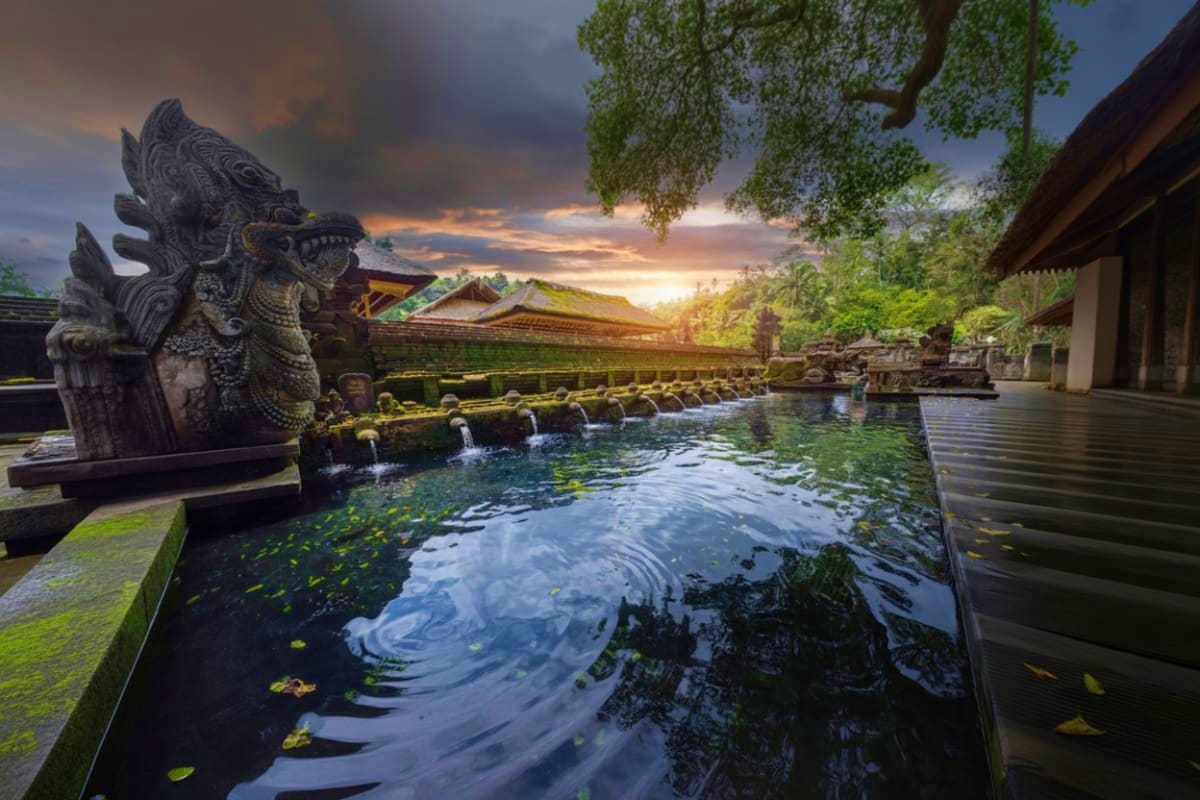 Ancient stone dragon statue beside sacred pool with rippling water at Tirta Empul Temple during sunset, lush greenery and tra