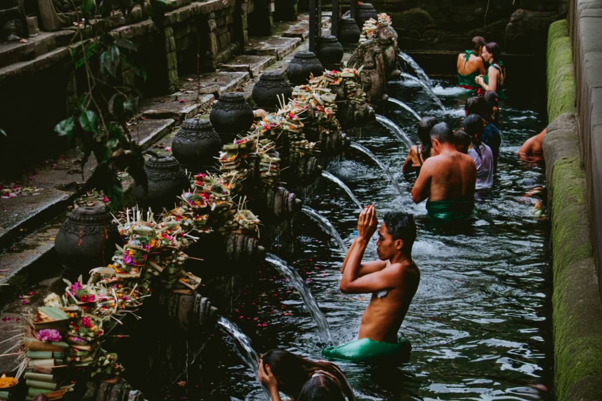 Visitors bathing in sacred spring water channels at Tirta Empul temple in Bali, with ornate stone spouts and colorful flower 