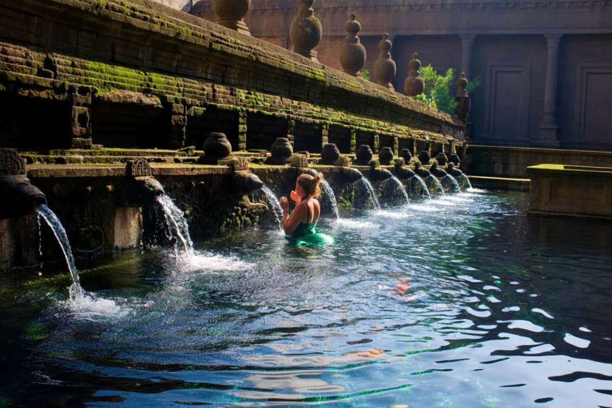 Person bathing in sacred spring water pool at Tirta Empul temple with moss-covered stone water spouts and ancient architectur