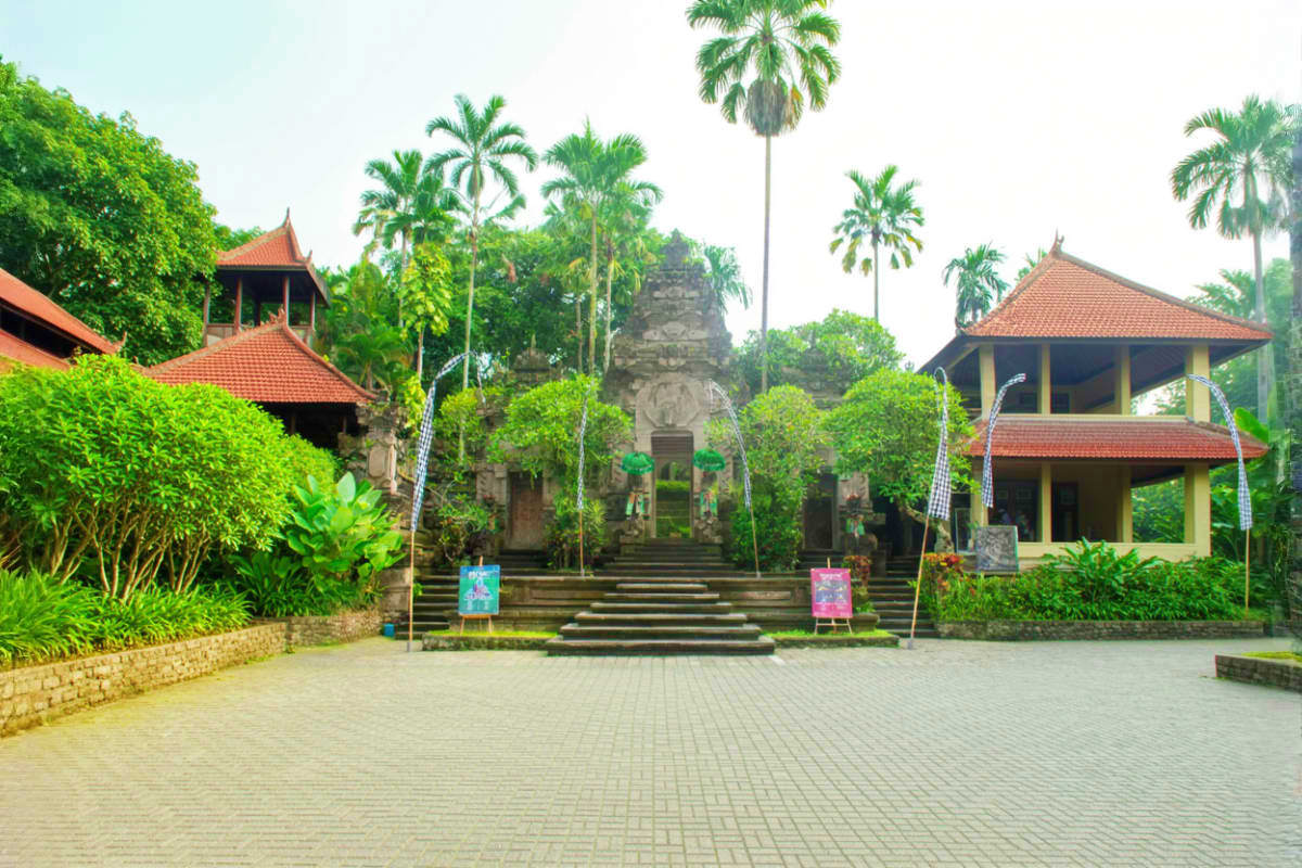 Balinese temple courtyard with stone gate, red-roofed pavilions, palm trees, and manicured gardens at Museum Puri Lukisan