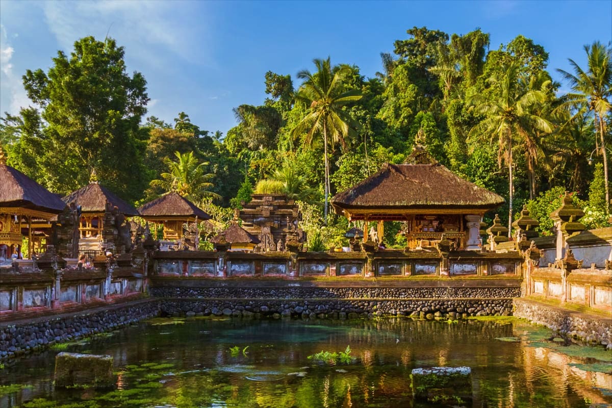 Traditional Balinese temple complex with thatched-roof structures reflected in clear water, surrounded by lush tropical veget