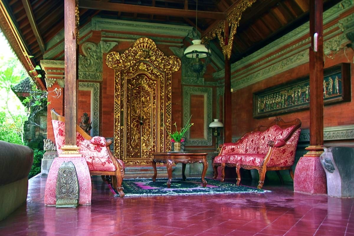 Ornate golden door and red velvet chairs inside traditional Balinese palace hall with decorative wooden beams and carved pane