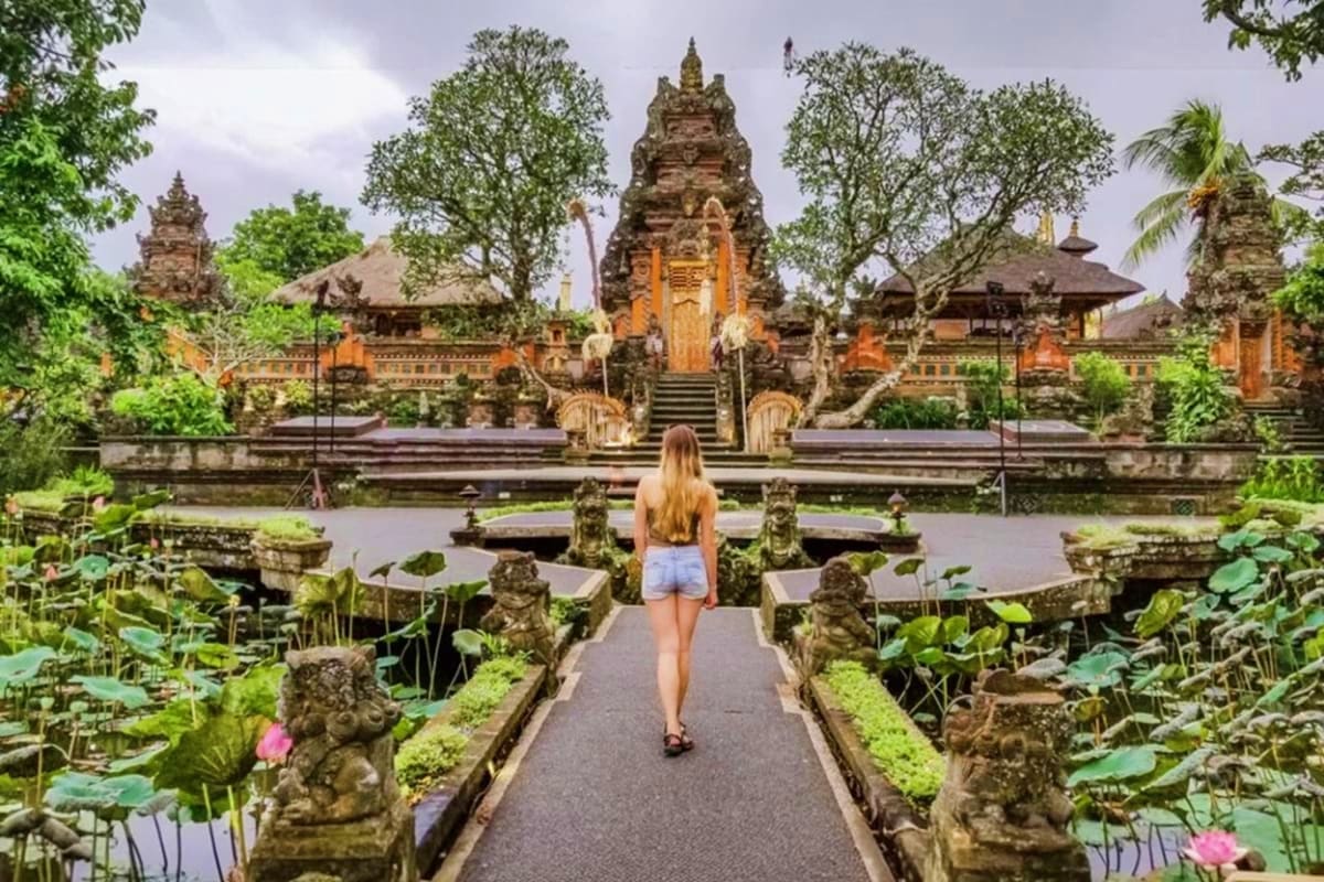 Woman walking on pathway toward Besakih Temple with ornate stone gates, lotus ponds, and traditional Balinese architecture su