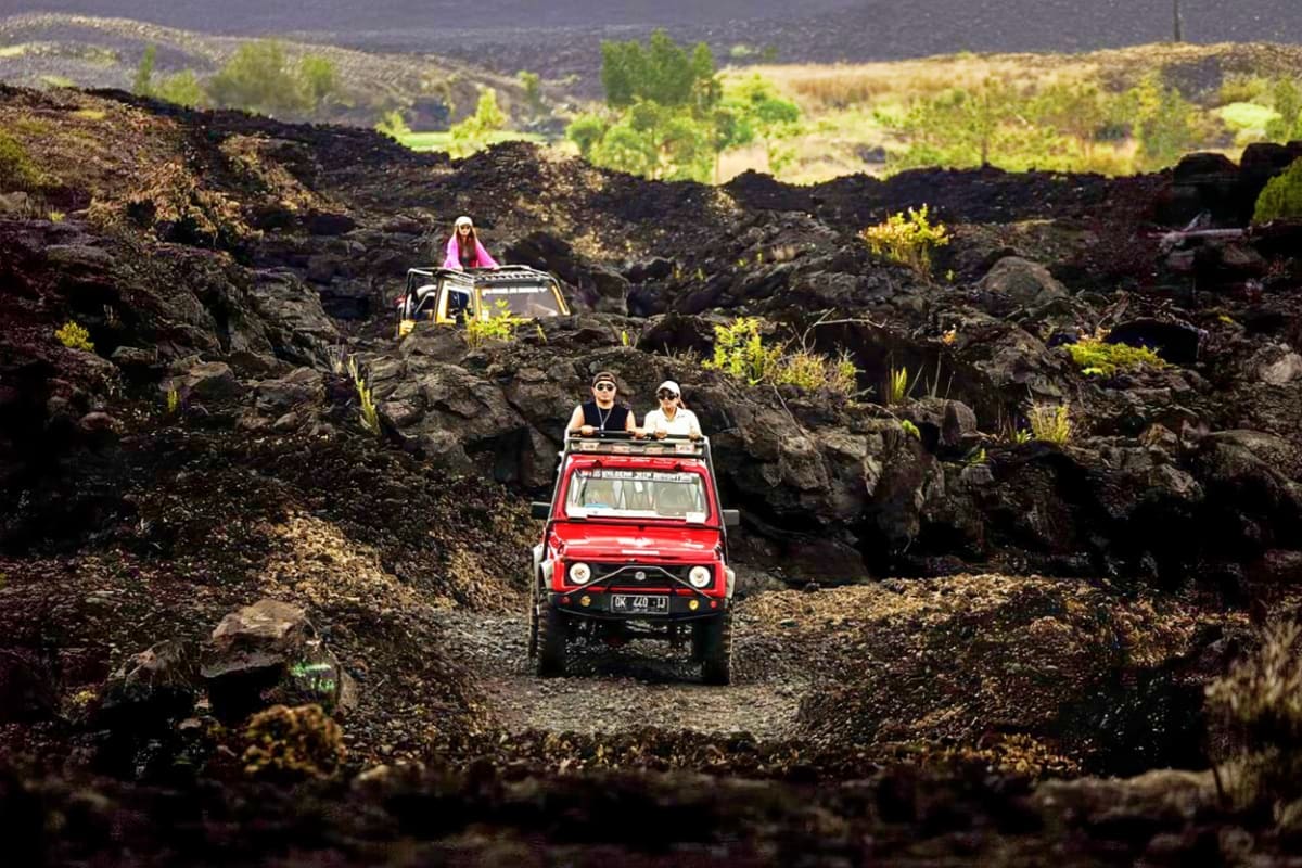 Tourists riding in red open-top jeeps navigating through rocky volcanic terrain with lush green valleys visible in the backgr
