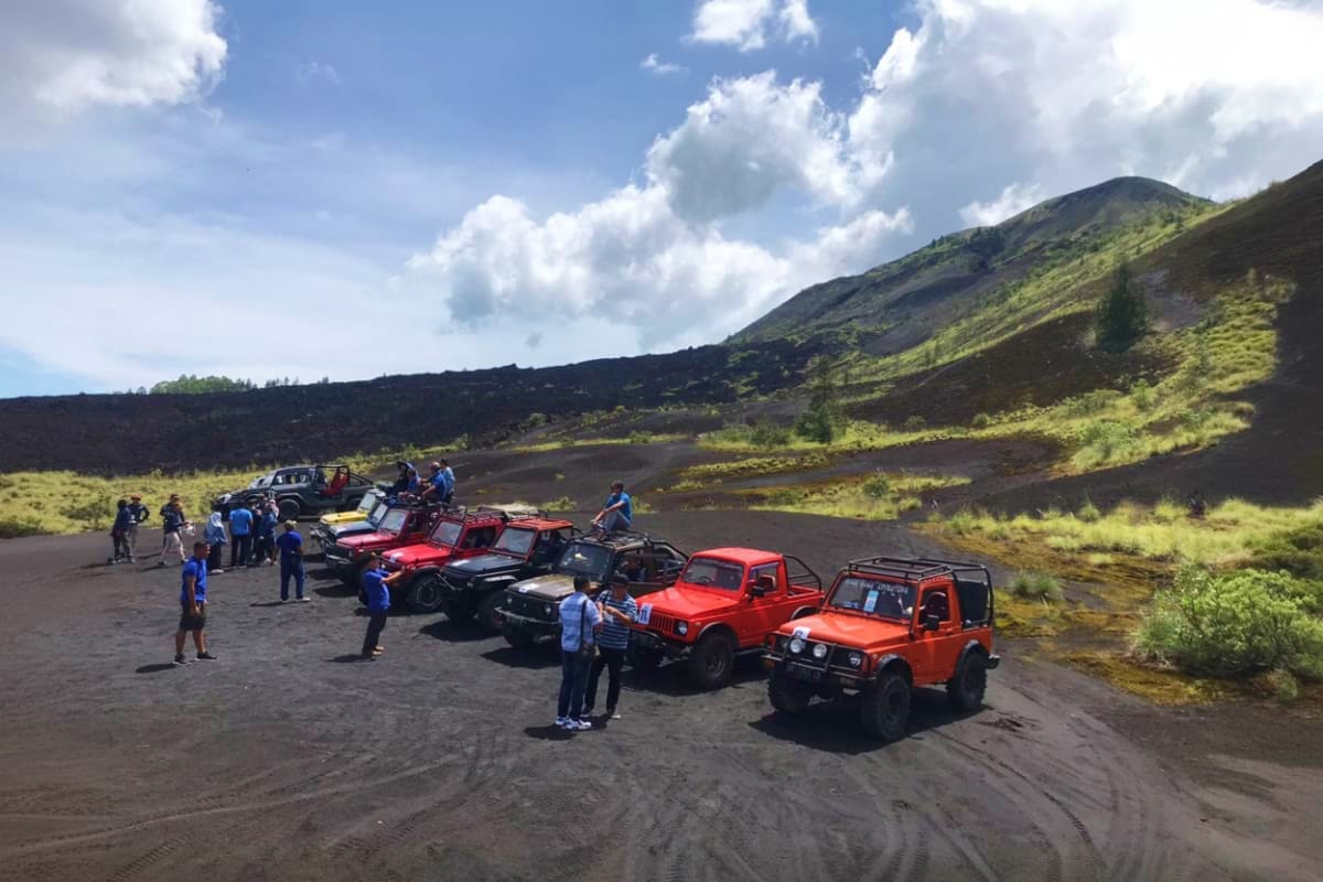 Group of tourists standing near red and black jeeps on black volcanic sand with Mount Batur and green hillside in background