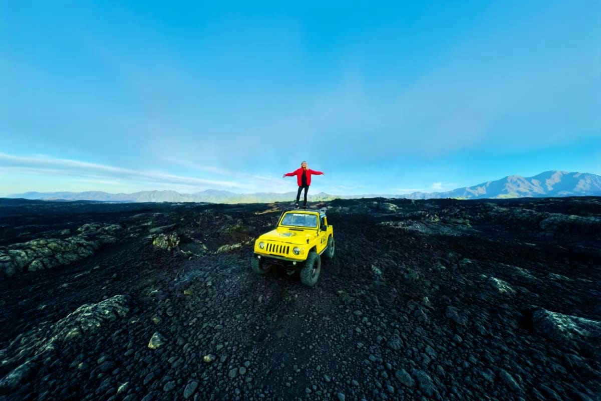 Person in red jacket standing on yellow Jeep on volcanic black lava field with mountains in background under blue sky