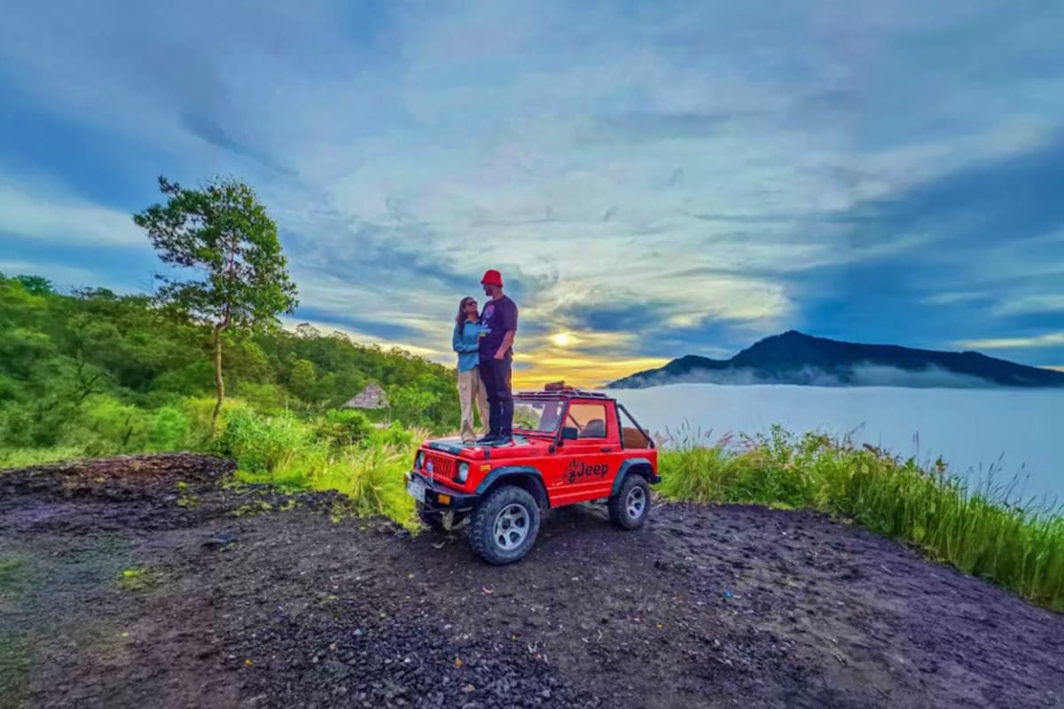 Couple standing on red Jeep overlooking Mount Batur lake at sunset with forested hillside and dramatic sky