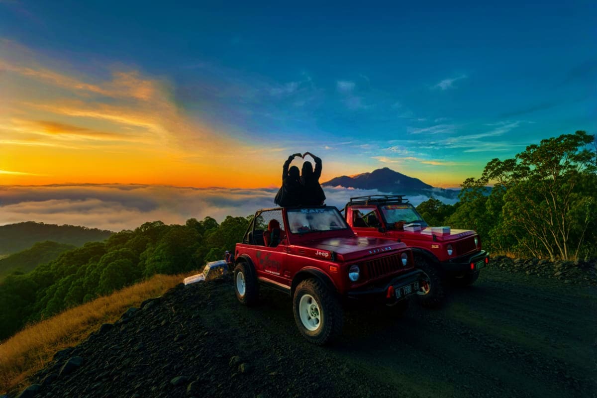 Couple making heart shape with hands on red Jeep at Mount Batur sunrise with volcanic mountain and cloud sea in background