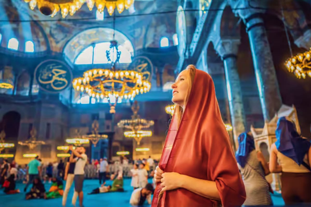 Woman in red headscarf gazing upward inside ornate mosque with blue walls, golden chandeliers, and domed ceiling decorated wi