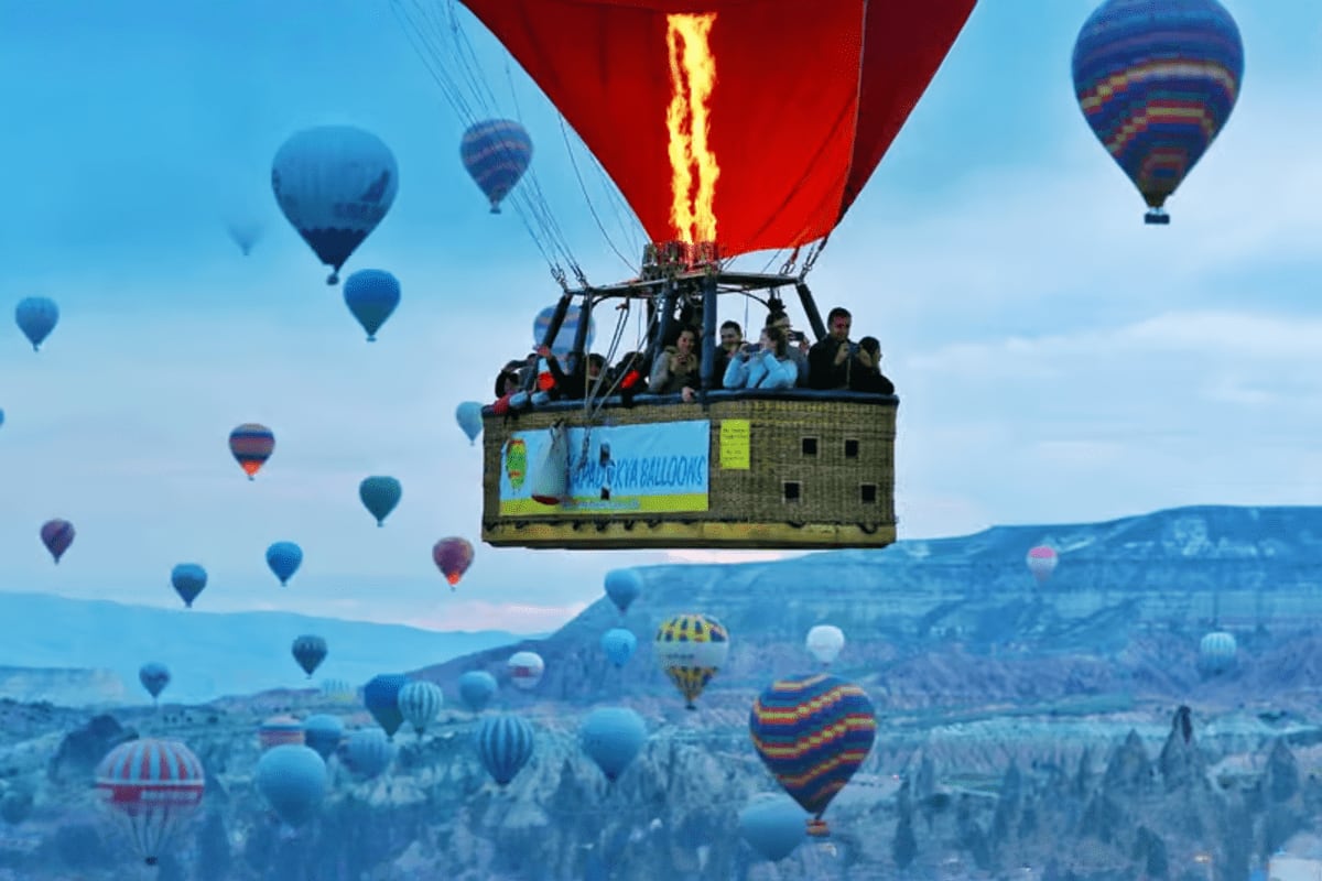 Hot air balloon with red envelope and flame burner above Cappadocia's fairy chimney landscape with dozens of other colorful b