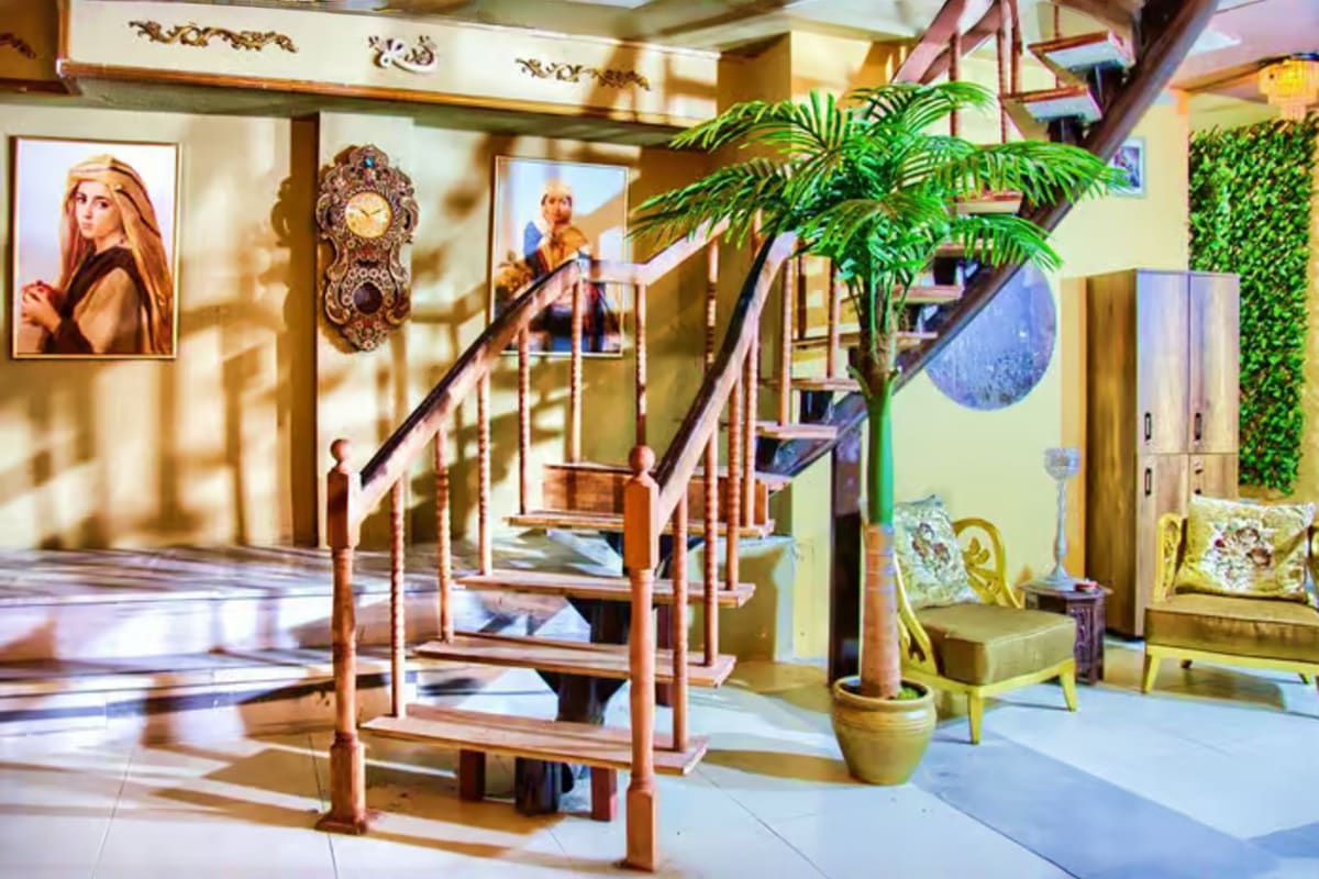Elegant hotel lobby with wooden staircase, ornate clock, framed portraits, potted plants, and decorative furnishings
