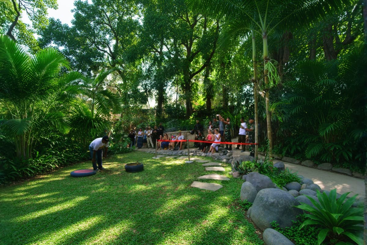 Outdoor garden area at Bali Reptile Park with visitors seated under red ribbon barrier, surrounded by palm trees and tropical