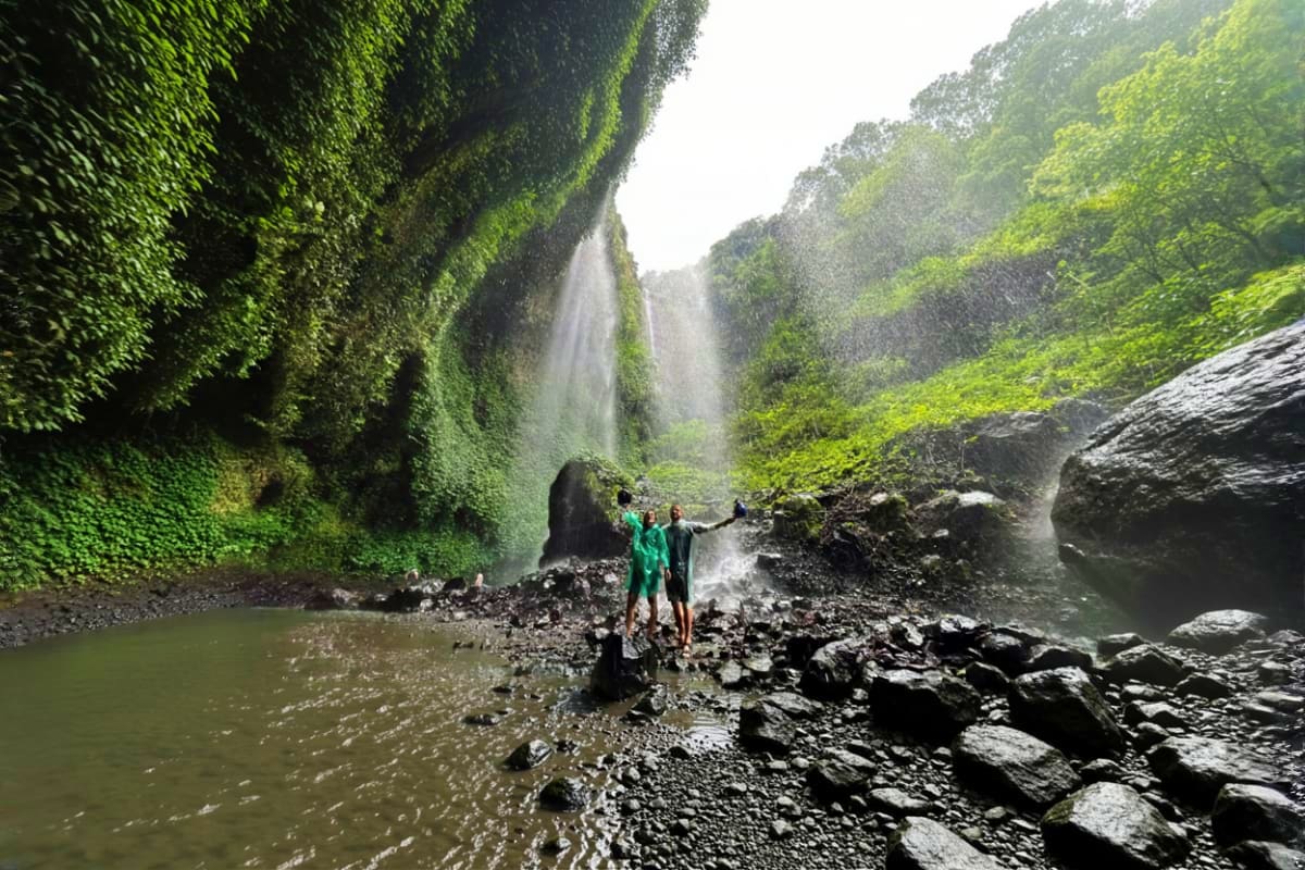 Two hikers standing on rocky terrain beneath a cascading waterfall surrounded by lush green vegetation and moss-covered canyo