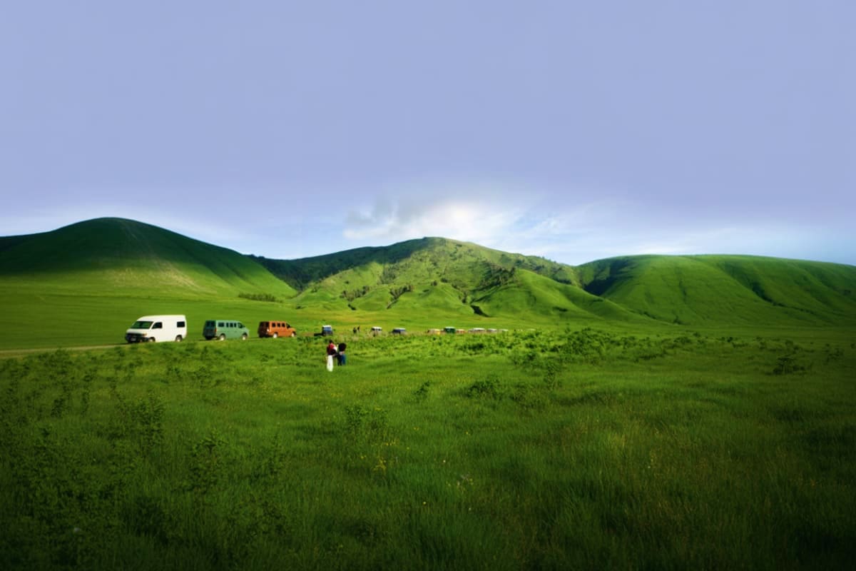 Vehicles parked in green savannah grassland with rolling hills and mountains in background during Mount Bromo tour