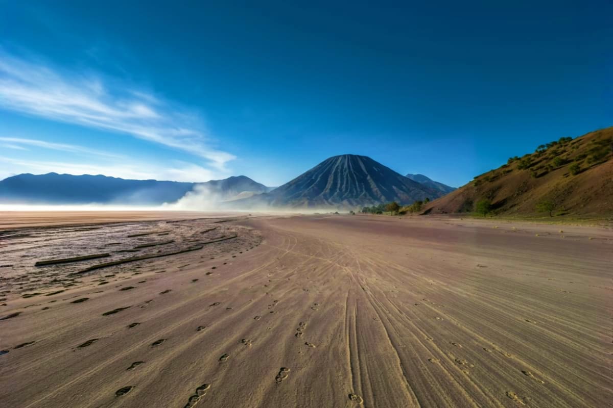 Mount Bromo volcanic cone with smoke rising above sand plain at sunrise, footprints visible on sandy beach foreground