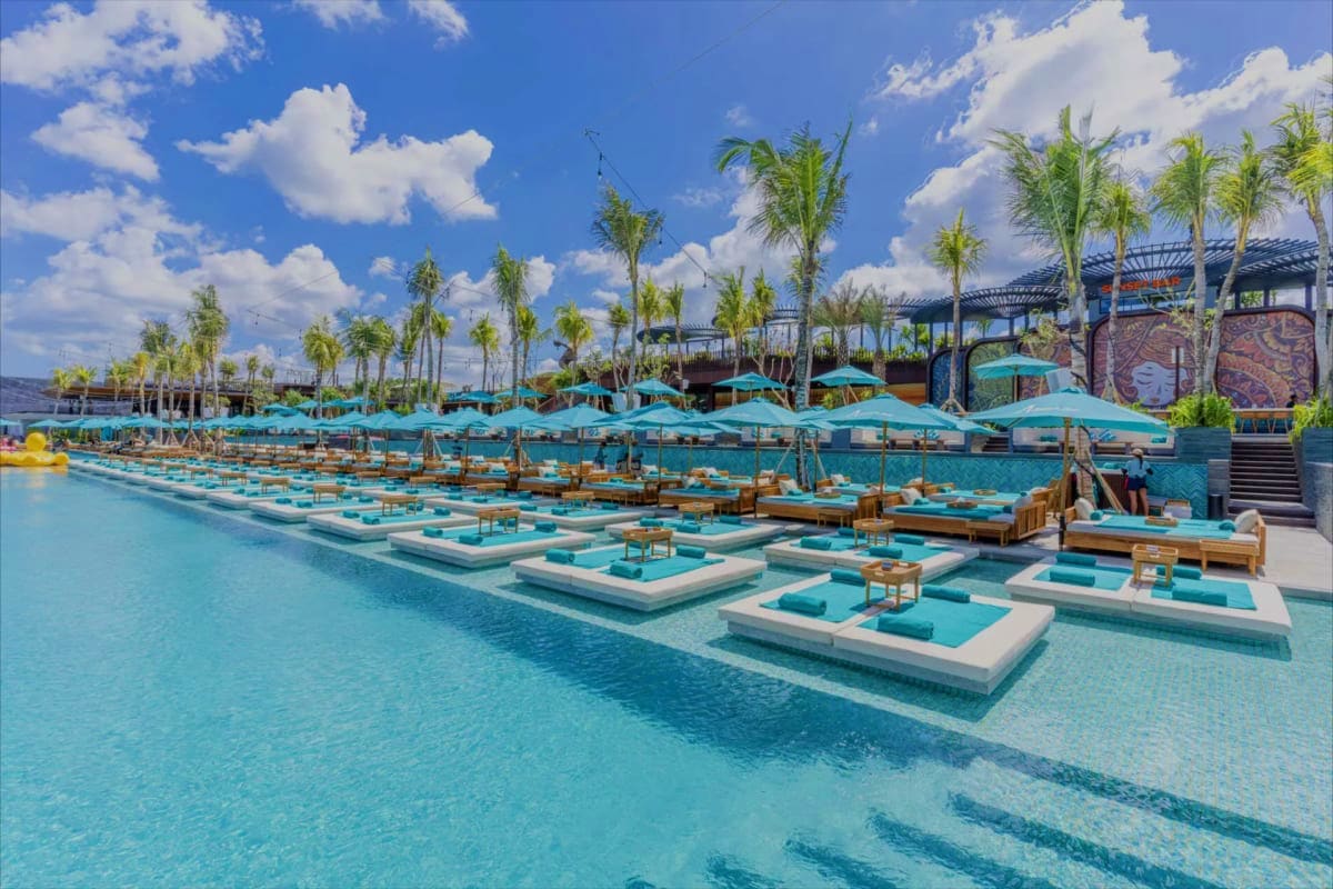 Luxury beach club with turquoise water, white sunbed platforms, teal umbrellas, palm trees, and modern architecture under blu