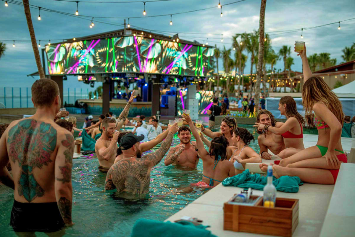 People swimming and socializing at Atlas Beach Club rooftop pool with neon sign, palm trees, and ocean view at dusk