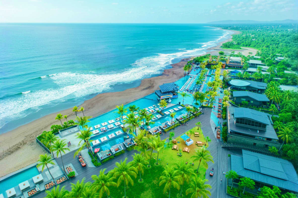 Aerial view of Atlas Beach Club with blue swimming pools, palm trees, loungers, and dark sand beach with ocean waves