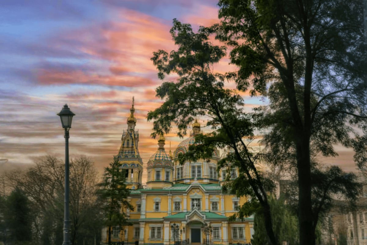 Orthodox cathedral with golden domes and yellow facade framed by trees at sunset in Almaty