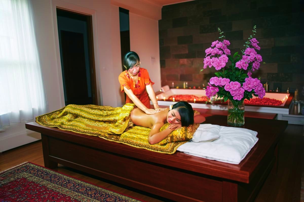 Thai massage therapist in orange uniform performing back massage on client wrapped in gold fabric at luxury spa with purple f