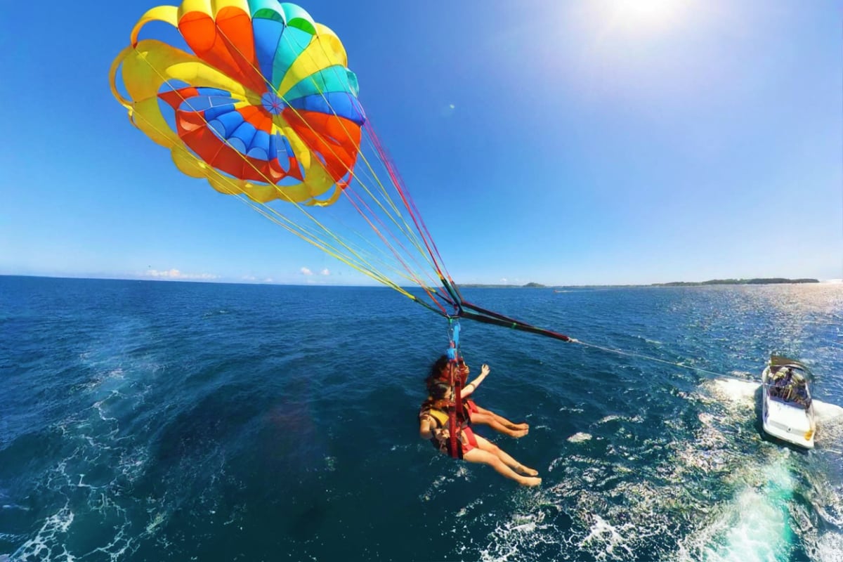 Two people parasailing behind a boat over turquoise ocean waters with colorful parachute canopy above, tropical coastline vis