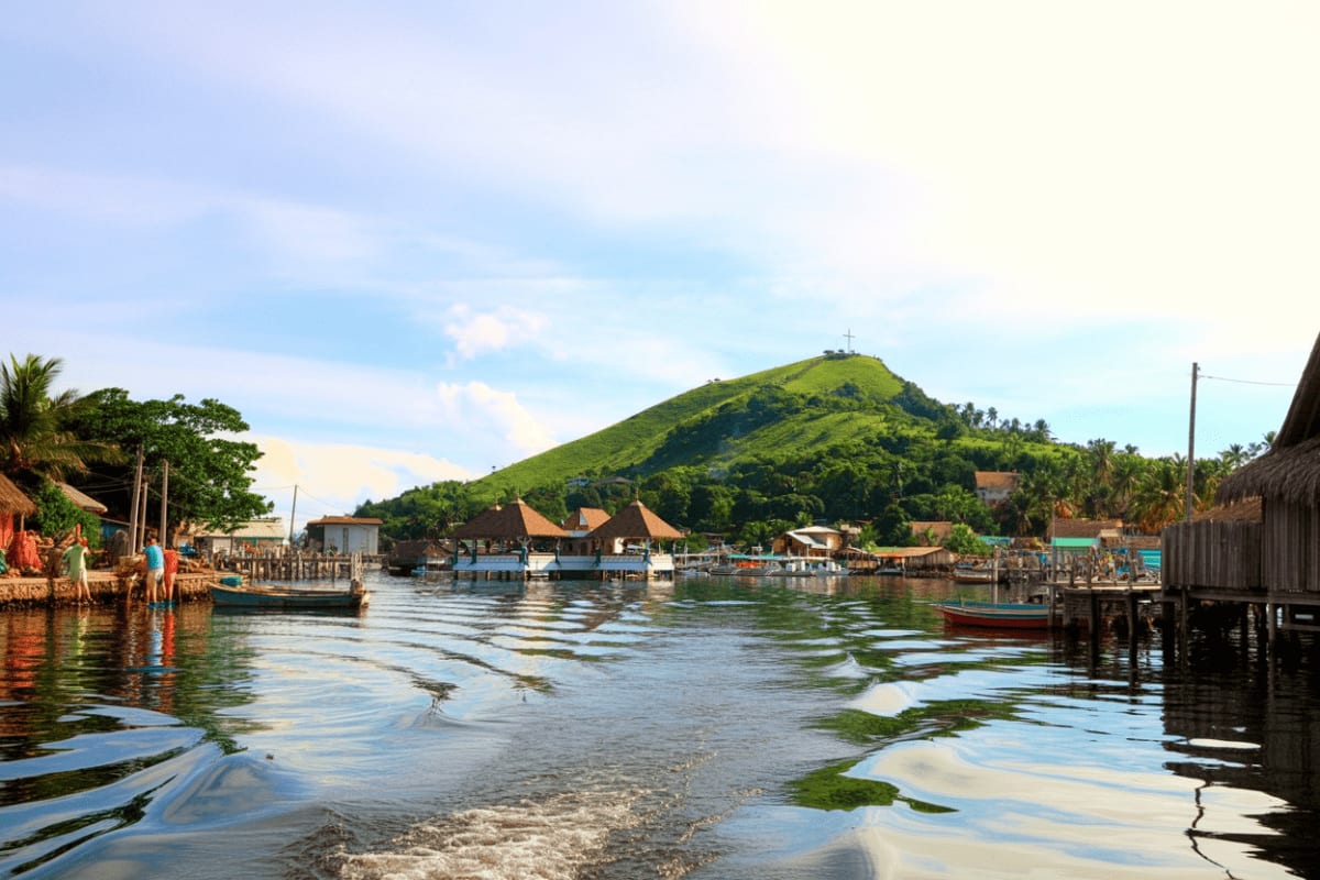 Coron town waterfront with traditional wooden houses on stilts, fishing boats, and green volcanic mountain with cross in back