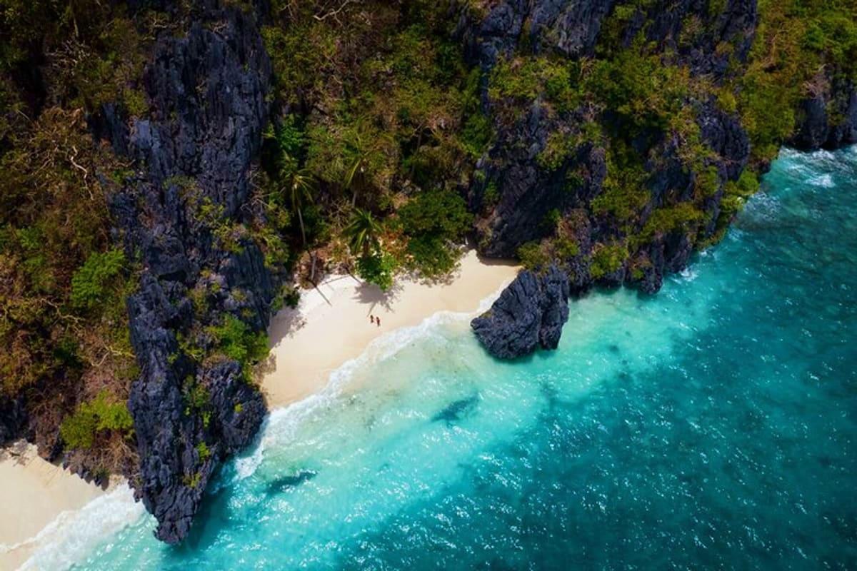 Aerial view of a secluded beach cove with turquoise waters, white sand, and towering limestone cliffs covered in green vegeta
