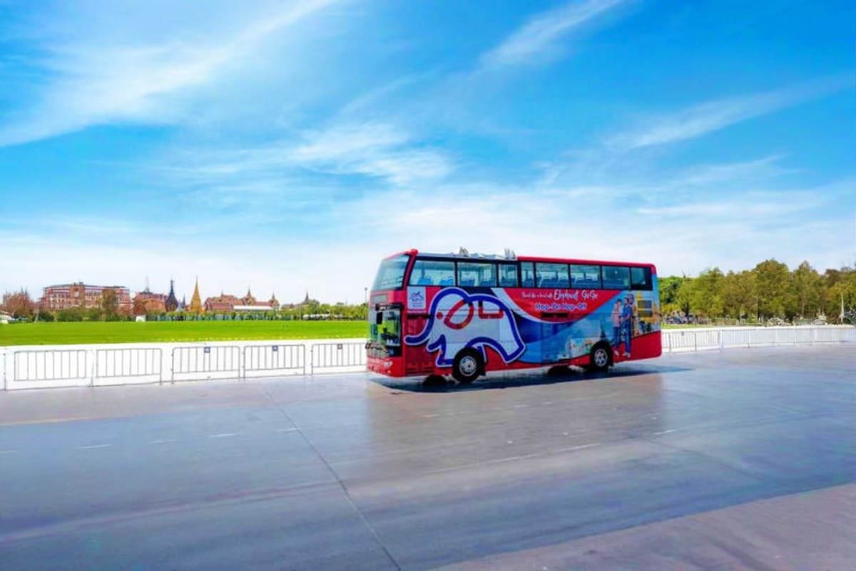 Red and blue double-decker tour bus parked on a waterfront promenade with city skyline and white railings in background under