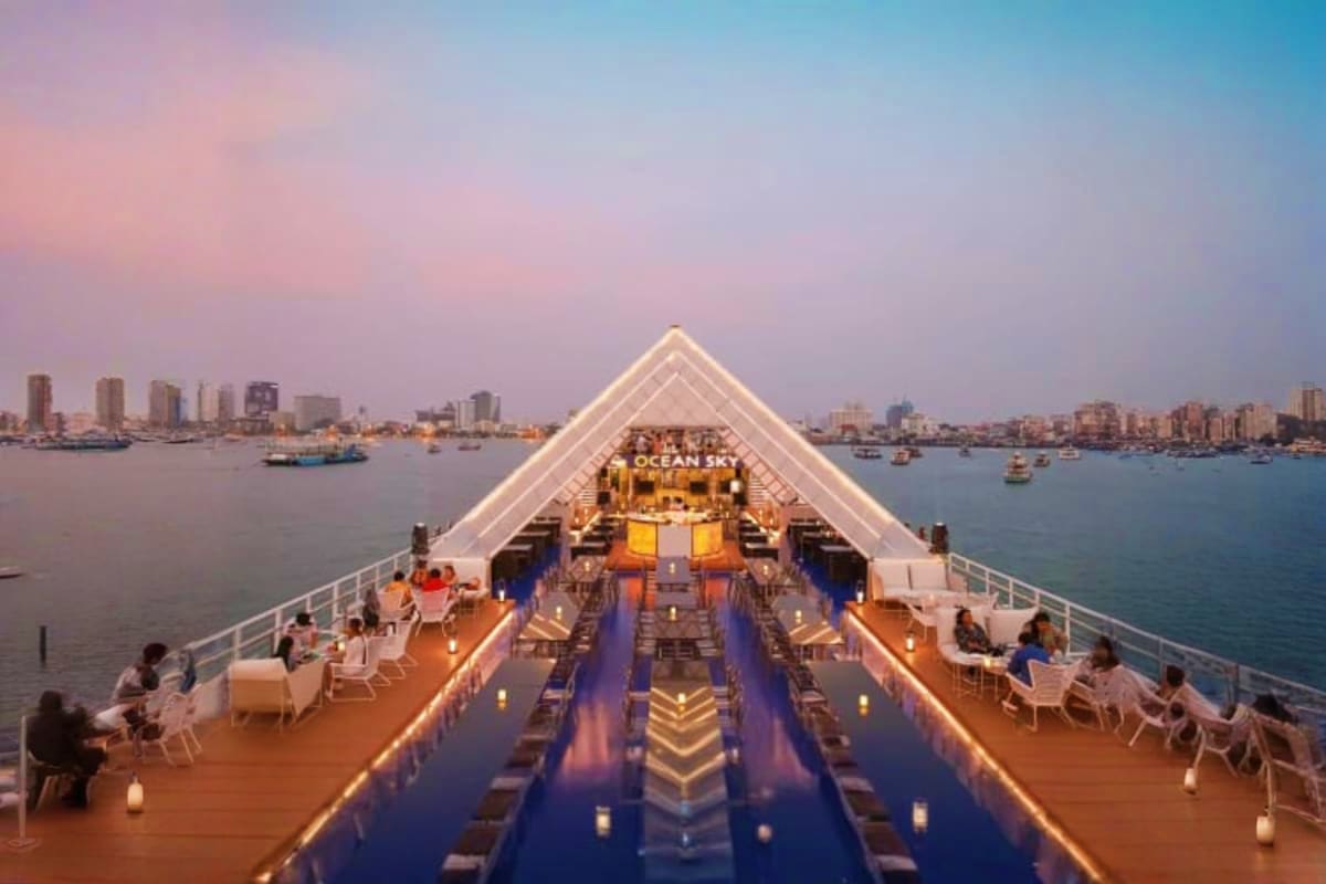Elevated view of illuminated cruise ship deck with passengers dining at sunset, city skyline and harbor in background