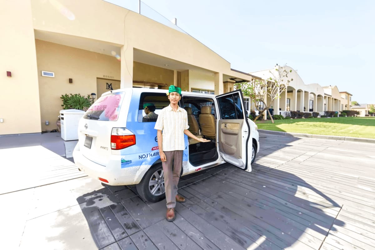 Man standing beside white and blue minivan with open side door at residential building, Bali airport transfer service