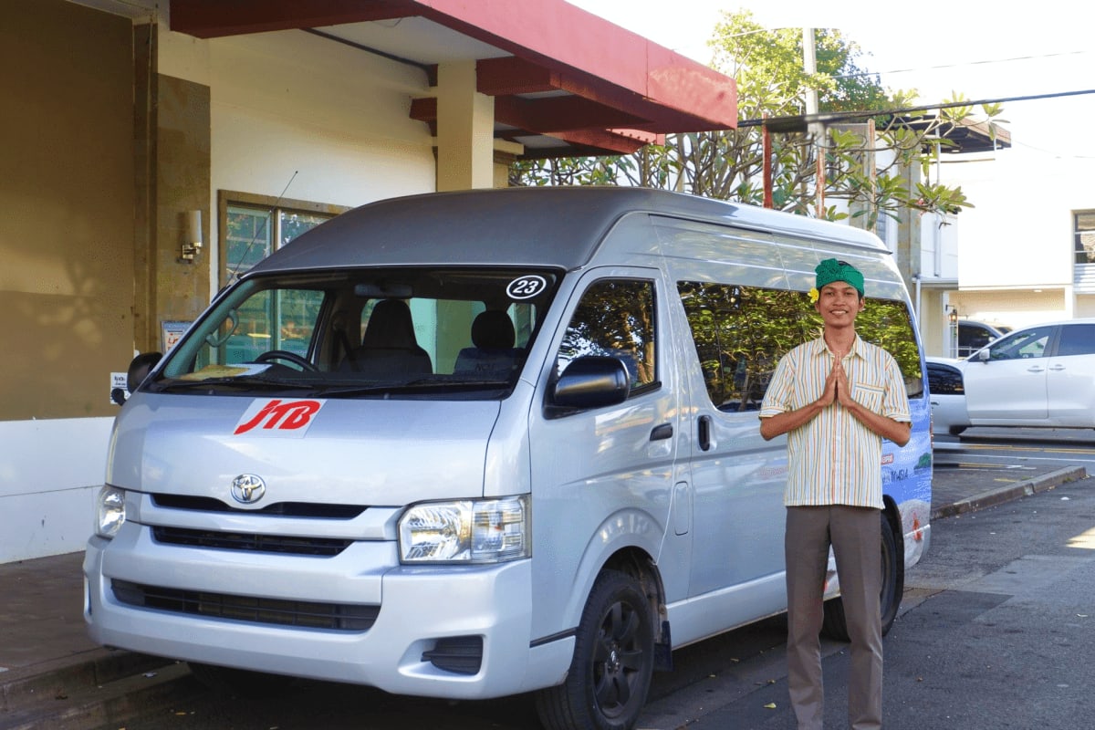 Male driver in green cap standing beside white Toyota van with "118" marking at Bali airport pickup location
