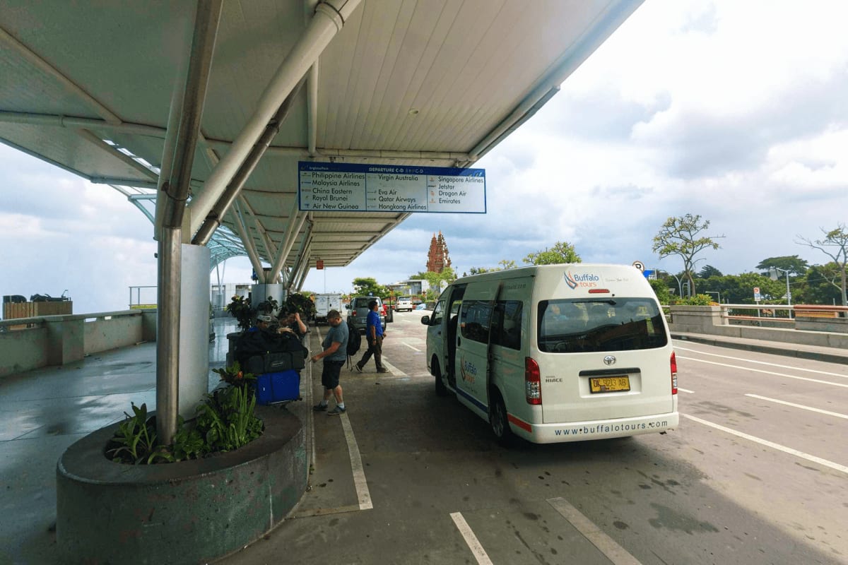 White shuttle van waiting at Bali airport departure area under covered structure with airline information sign visible