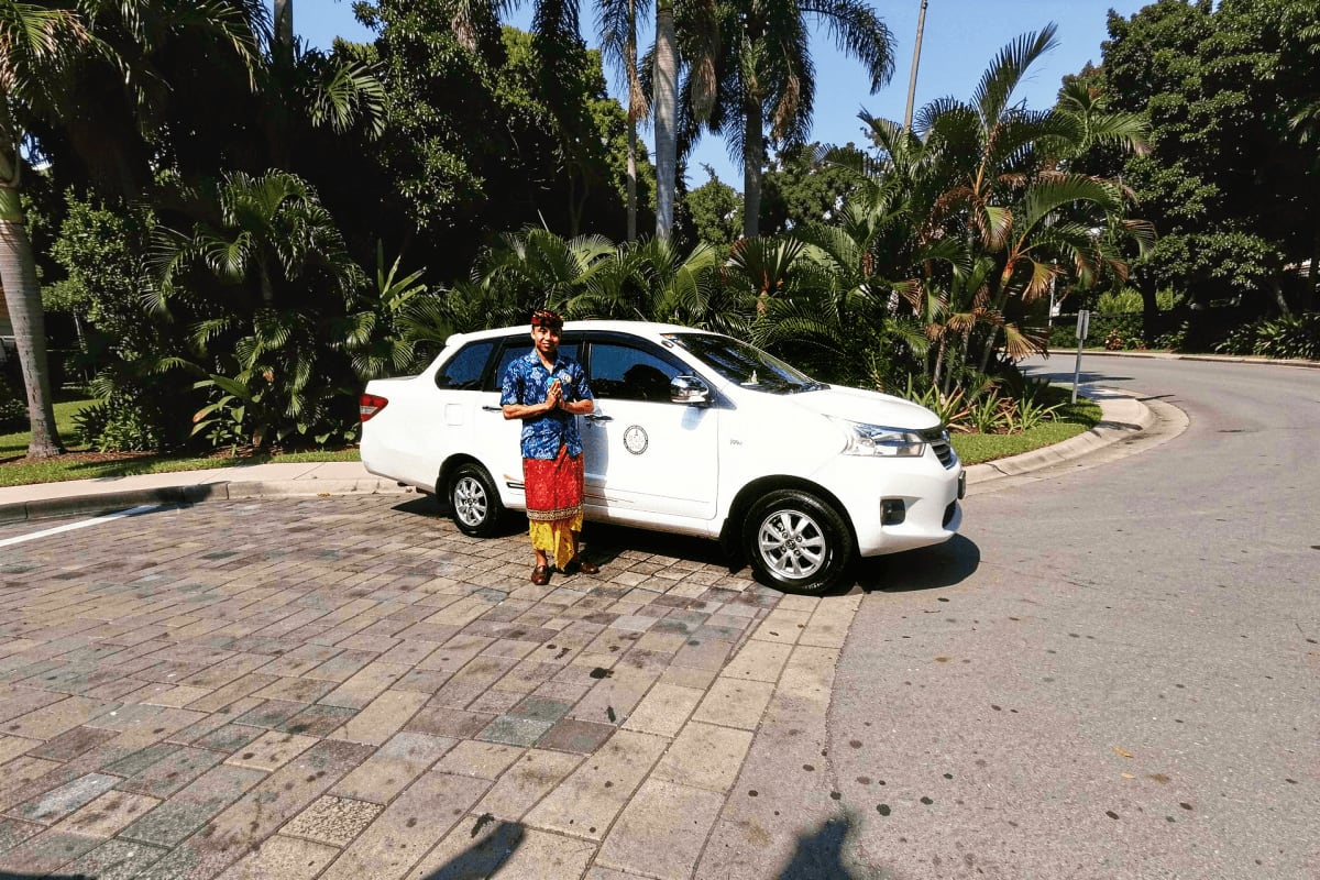Man standing beside white car in tropical palm tree-lined driveway for airport pickup service