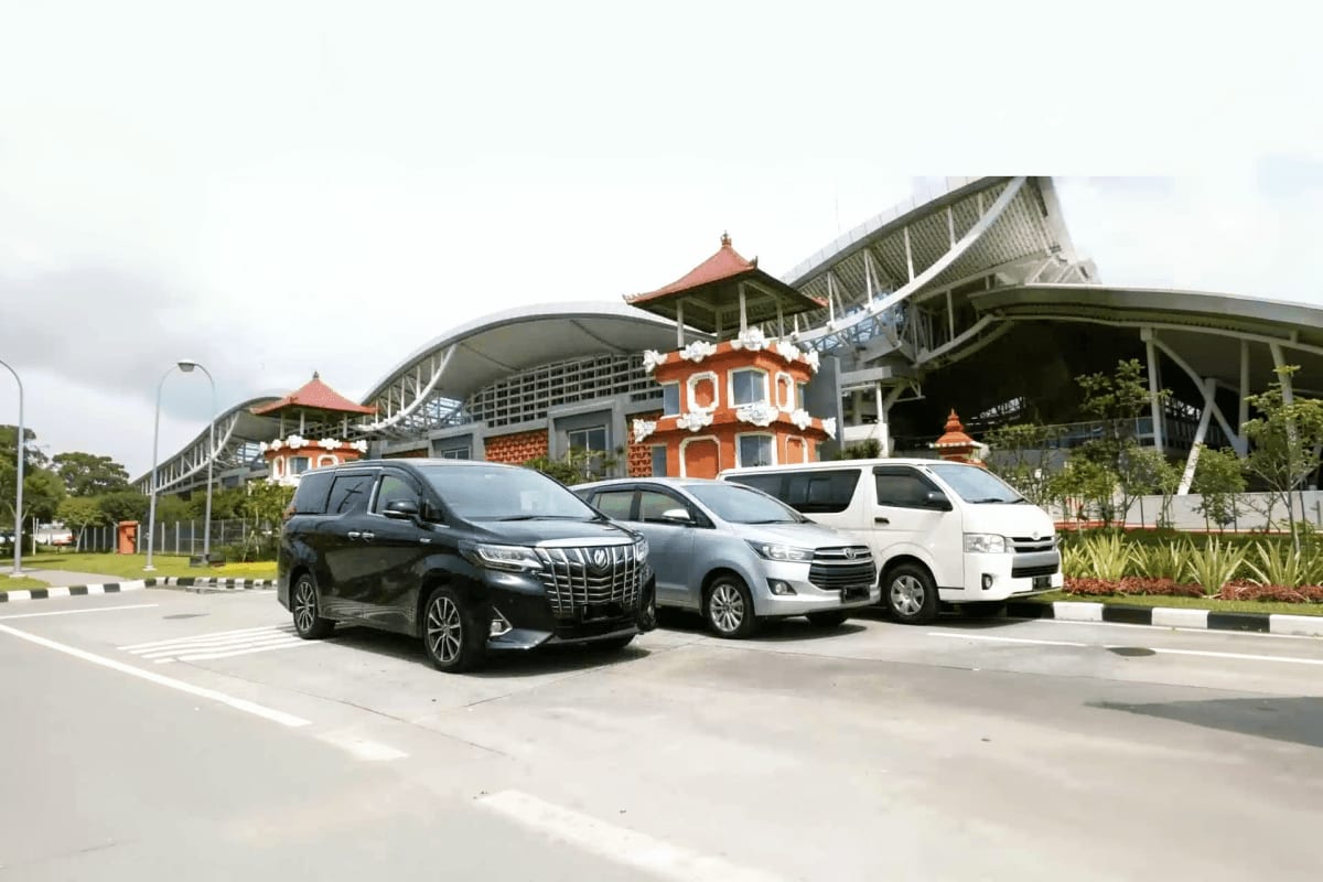 Three minivans parked at Bali Airport with traditional red-roofed architecture building in background