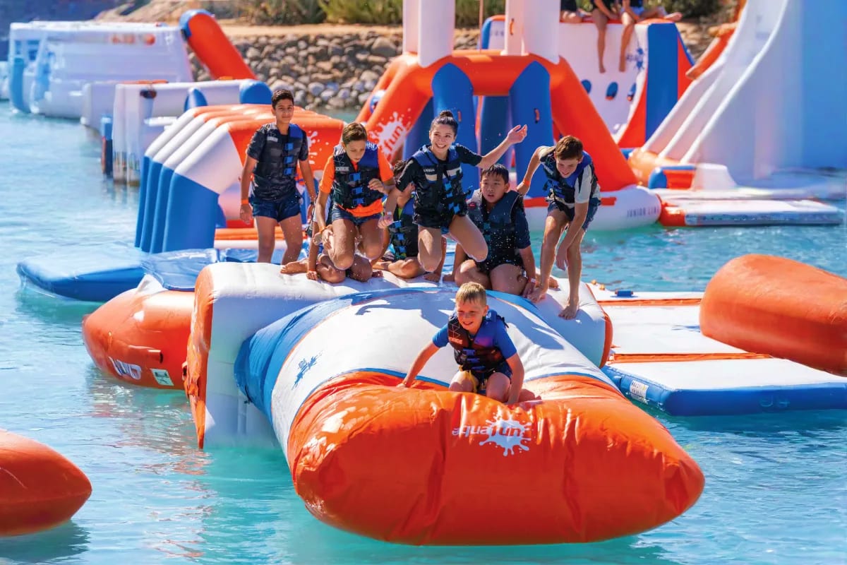 Children enjoying inflatable water obstacles and slides at an aquatic adventure park with blue and orange equipment in shallo