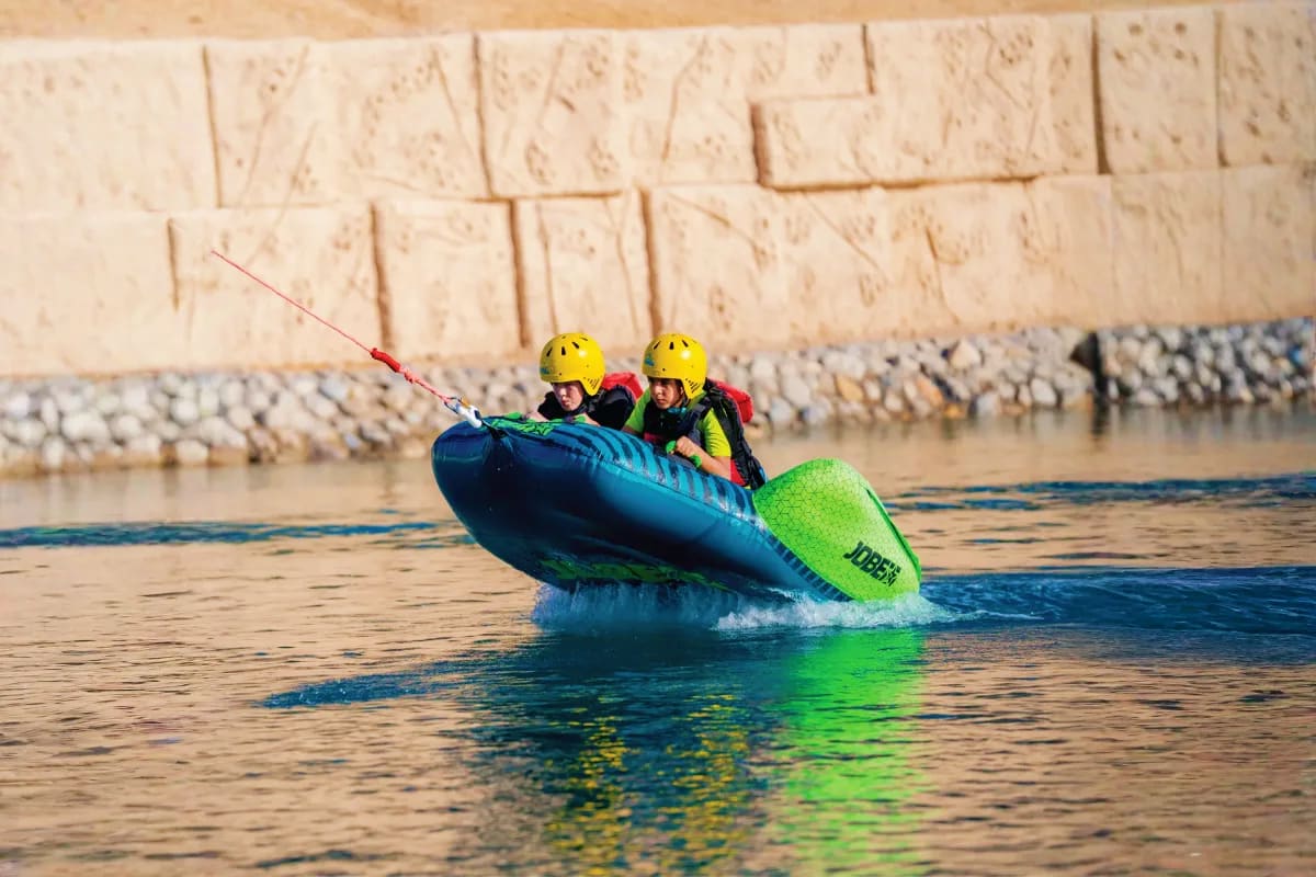 Two people in yellow helmets and life jackets riding in a blue and green inflatable boat on calm water near a stone barrier w
