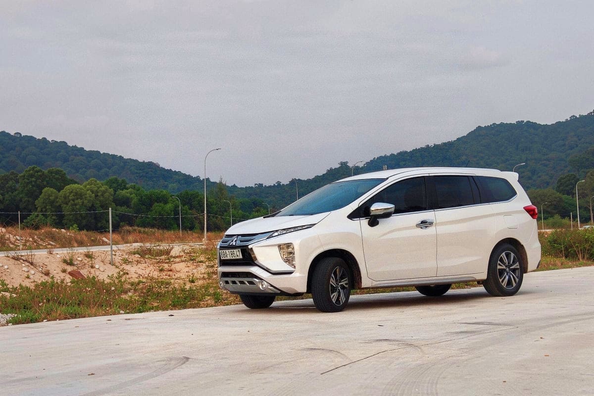 White Mitsubishi Expander MPV parked on concrete surface with forested mountains in background under cloudy sky