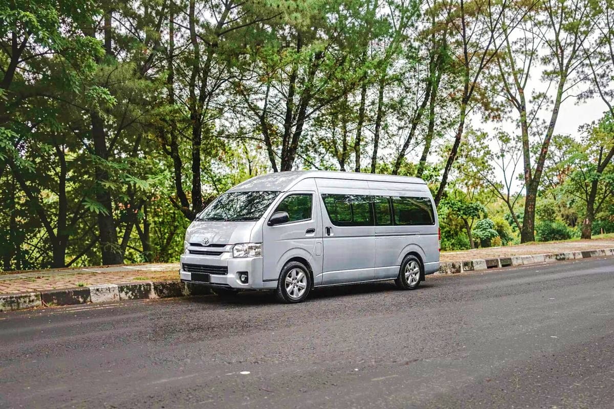 Silver Toyota minivan parked on asphalt road with dense green trees in background, available for private hire transport