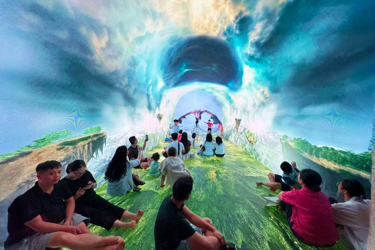 Visitors sitting on moss-covered ground inside an immersive digital art tunnel with glowing blue sky and nature projections o