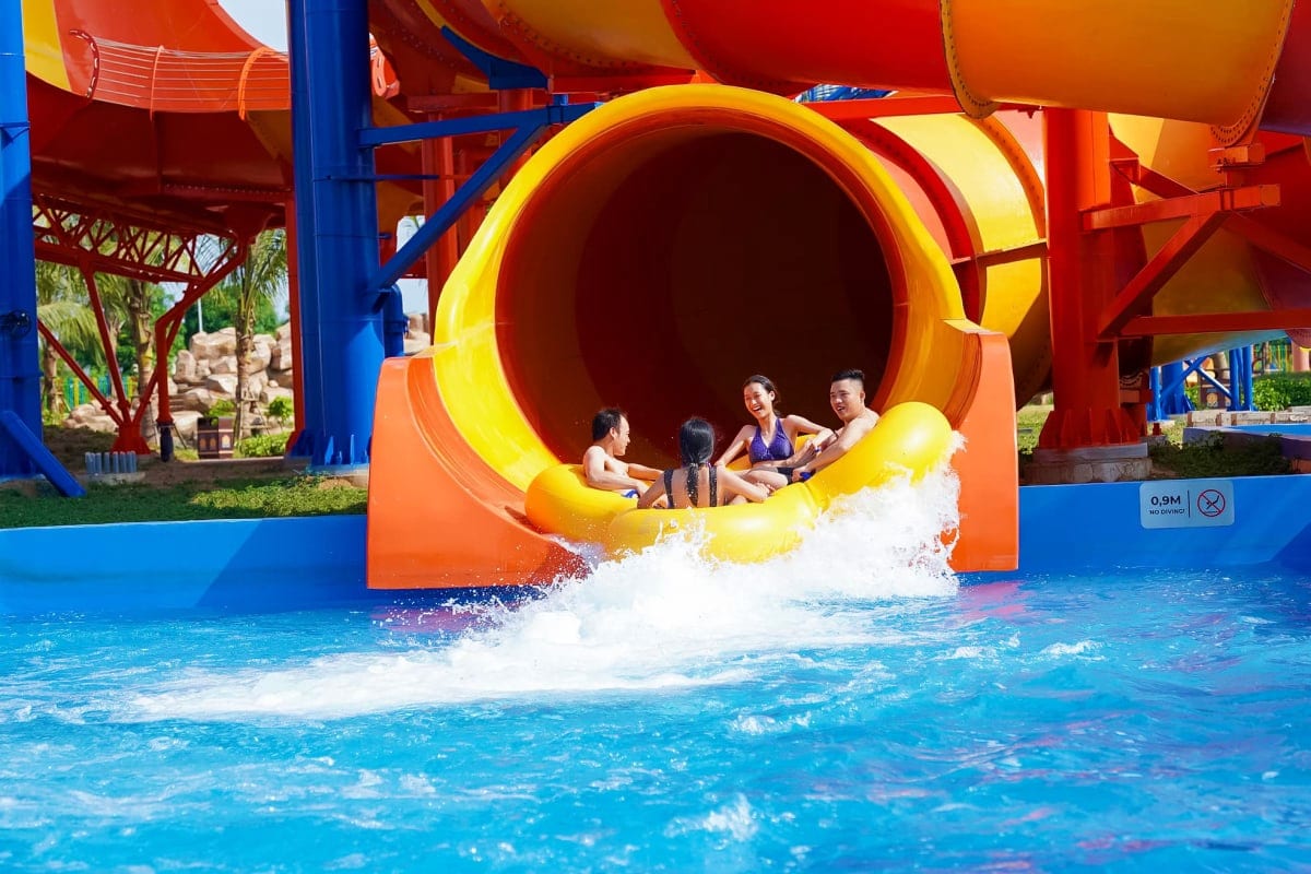 Children sliding down a large yellow and red tunnel water slide into a bright blue pool at Vinwonders Cua Hoi water park, cre