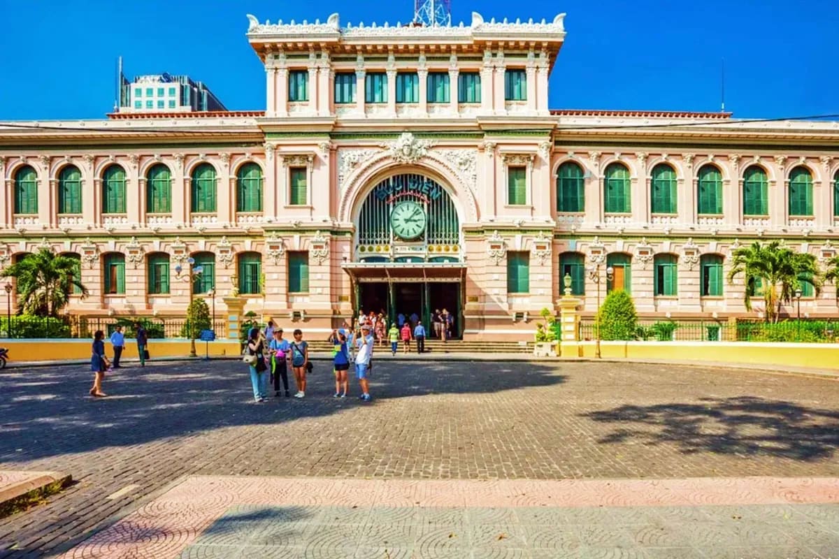 Colonial-era pink building with clock tower and green shutters, tourists gathered in courtyard under blue sky, Ho Chi Minh Ci