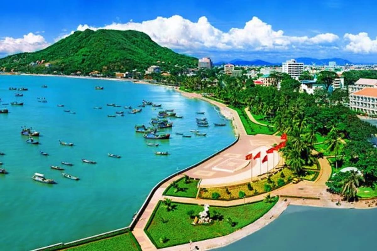 Aerial view of Vung Tau coastal town with turquoise bay, fishing boats, green hillside, red flags on beach, and resort buildi