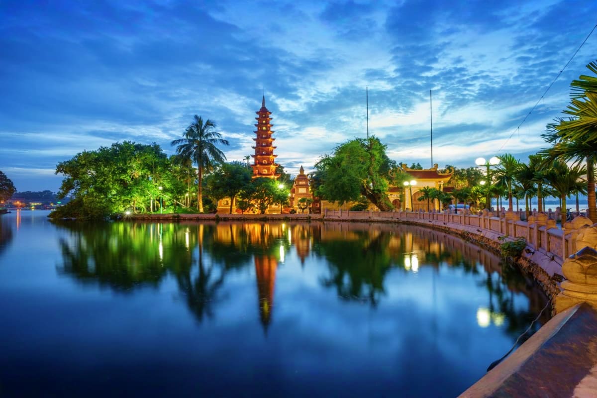 Tran Quoc Pagoda illuminated at dusk reflected in Hanoi's Red River with palm trees and traditional architecture