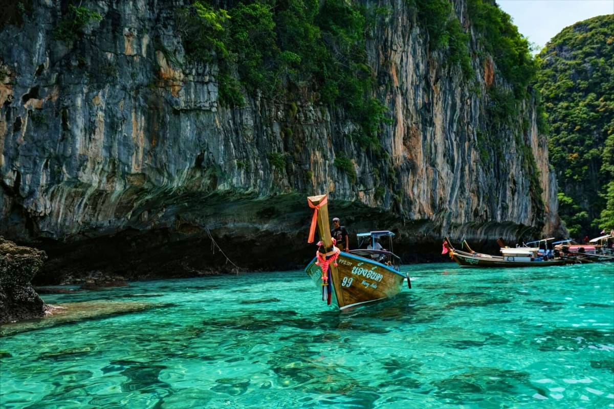 Traditional long tail boat anchored in crystal clear turquoise water beneath towering limestone cliffs in tropical bay