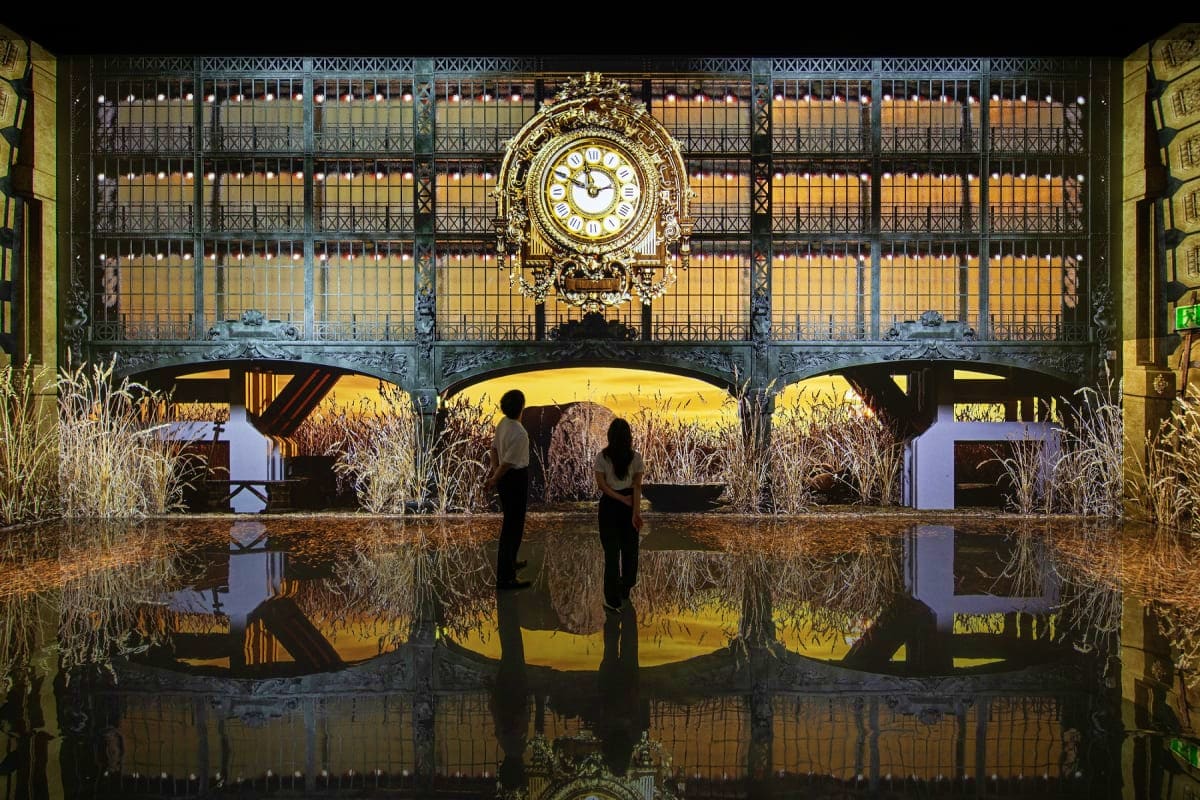 Two visitors standing before ornate wrought-iron gates with golden clock at Arte Museum Dubai, reflected in wet floor