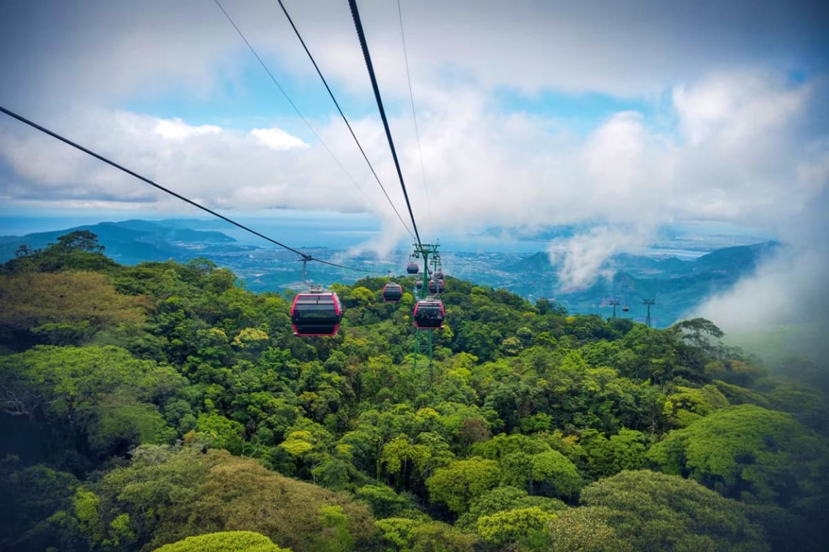 Cable cars with red cabins suspended above lush green forest canopy with mountains and blue sky in background at Ba Na Hills