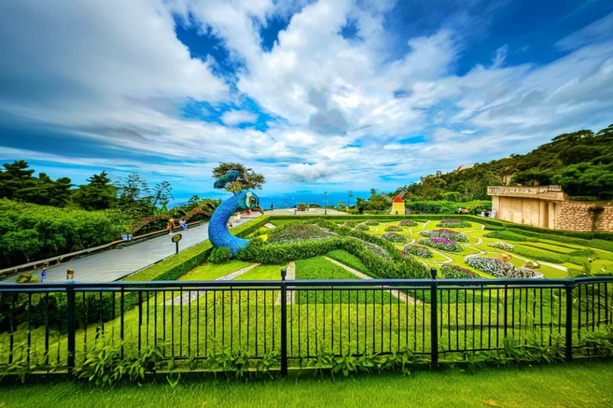 Panoramic view of Bana Hills garden with blue peacock sculpture, manicured lawns, and mountain landscape under blue sky with 
