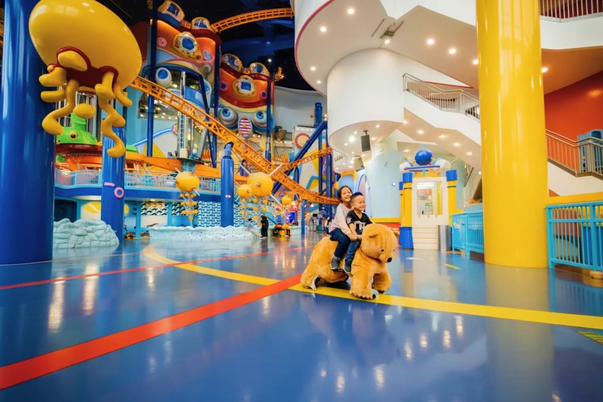 Children riding on a yellow plush dog attraction inside a colorful indoor theme park with blue, yellow and orange play struct