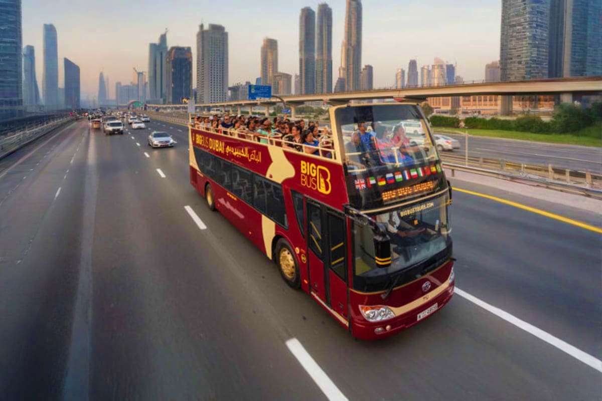 Red and yellow double-decker Big Bus tour bus driving on Dubai highway with modern skyscrapers and cityscape in background