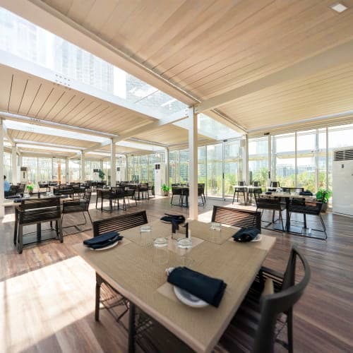 Modern open-plan restaurant with floor-to-ceiling windows overlooking city skyline, featuring dining tables, black pianos, an